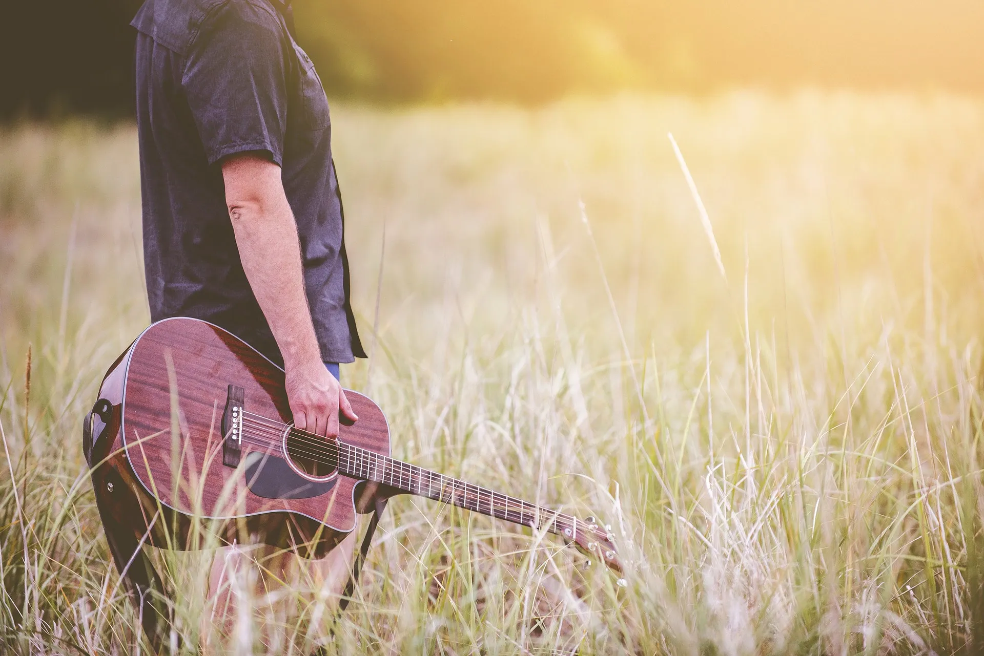 A musician holding an acoustic guitar in tall grass at sunset